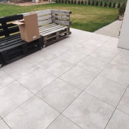 BUD CORP - Wyłożony jasnymi płytkami taras z narożną sofą z palet, jedna pomalowana na czarno na kółkach, druga naturalna, częściowo zdemontowana, z kartonem na siedzisku.