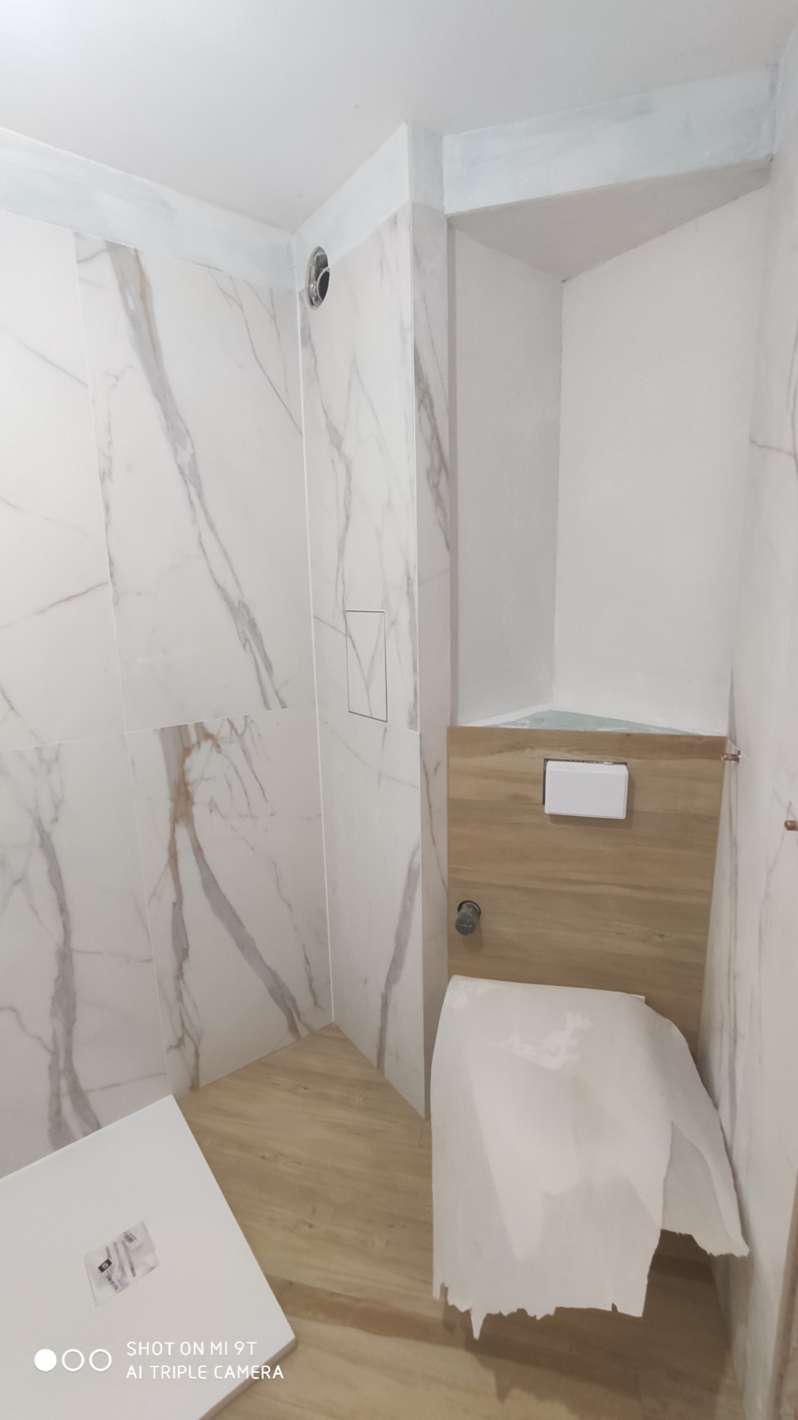 Wnętrze łazienki w trakcie wykańczania: marmurowe płytki na ścianach, drewnopodobna podłoga, zabudowa WC z drewnianymi panelami, częściowo zakryta toaleta, widoczny otwór wentylacyjny w suficie...