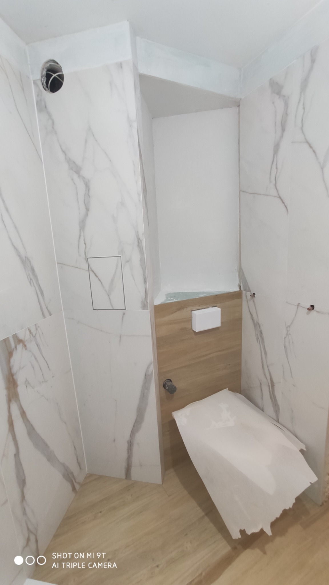 Łazienka w trakcie remontu: białe marmurowe płytki, drewnopodobna zabudowa, częściowo zakryta toaleta, widoczne ślady po pracach wykończeniowych.