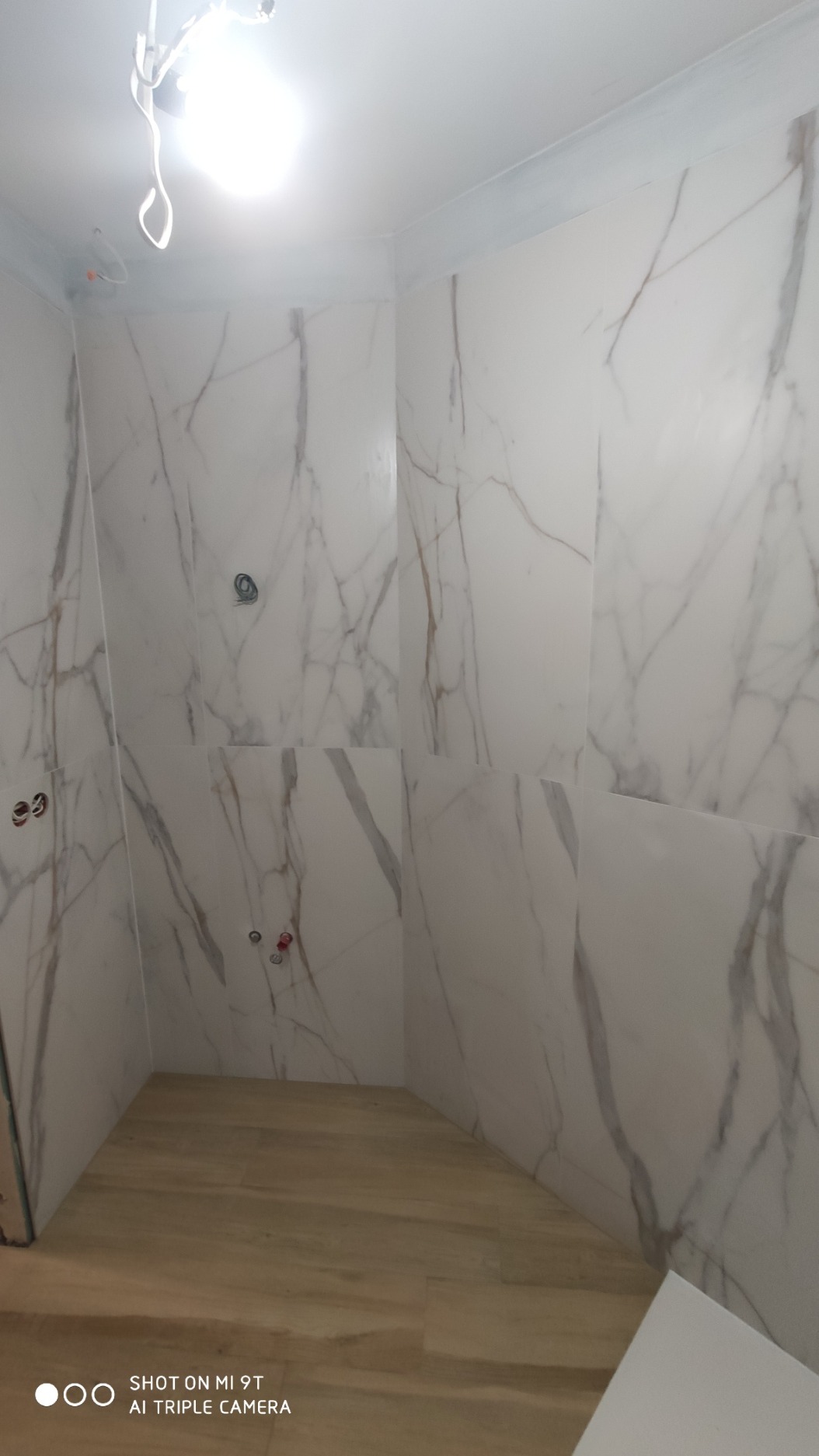 Świeżo wykafelkowana ściana łazienki z białymi płytkami imitującymi marmur, widoczne otwory na armaturę, drewnopodobna podłoga, surowe wykończenie sufitu.