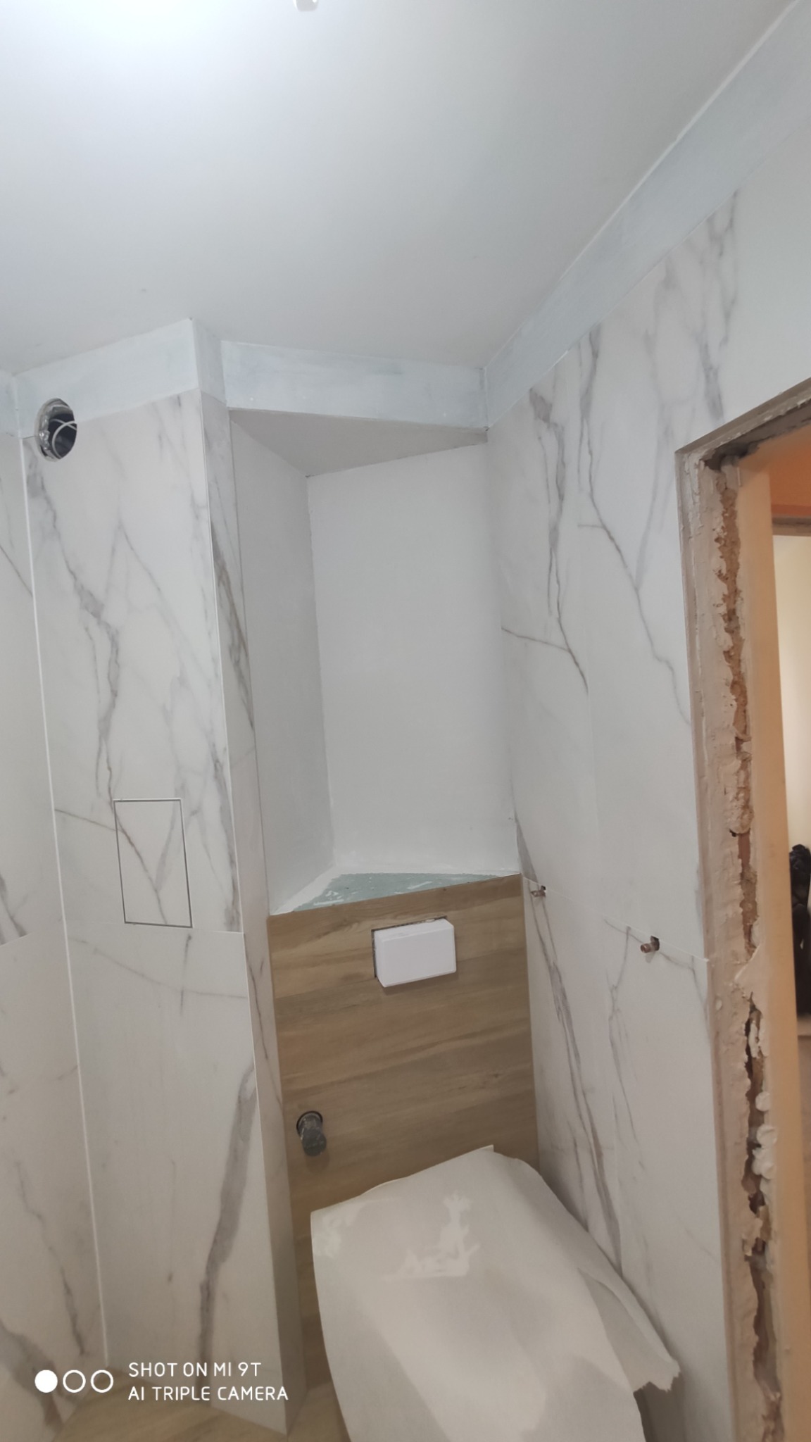 Łazienka w trakcie remontu: białe płytki imitujące marmur na ścianach, częściowo zabudowana toaleta podtynkowa z drewnianym panelem, widoczne ślady po pracach remontowych wokół futryny.