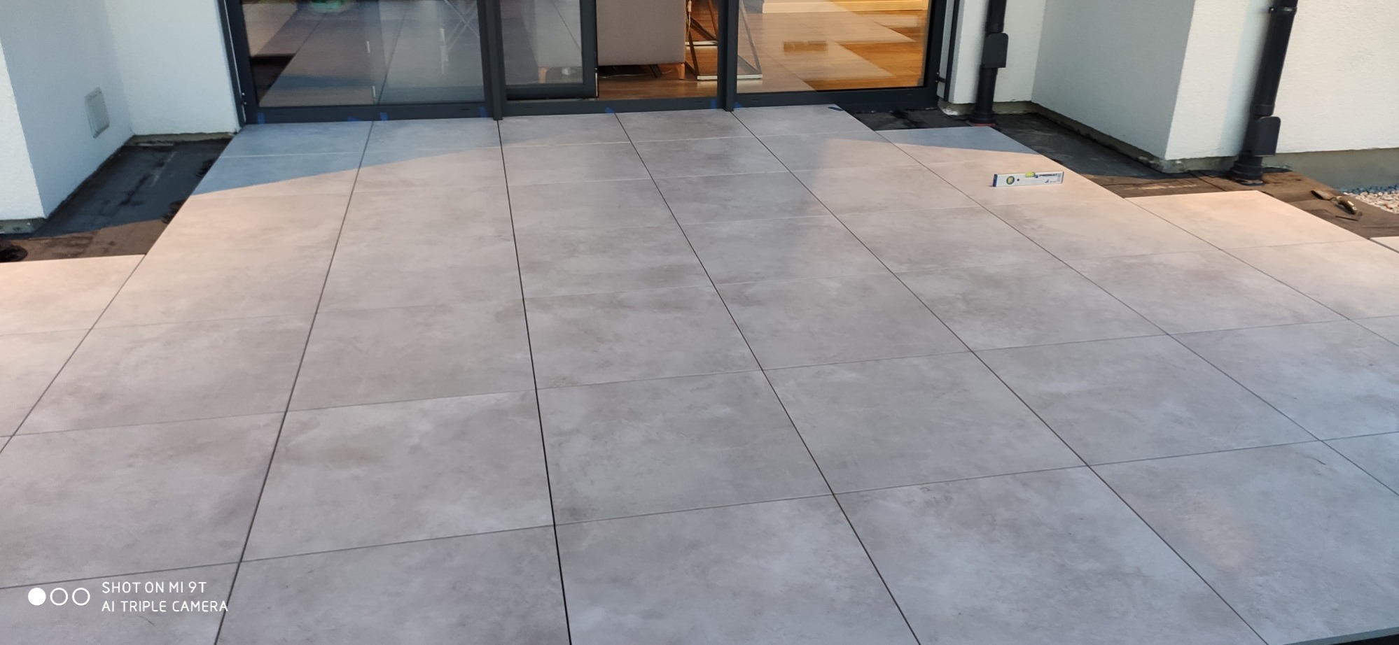 Jasnoszare płytki ułożone na zewnątrz, przy drzwiach tarasowych, poziomica leżąca obok, widoczny fragment izolacji.