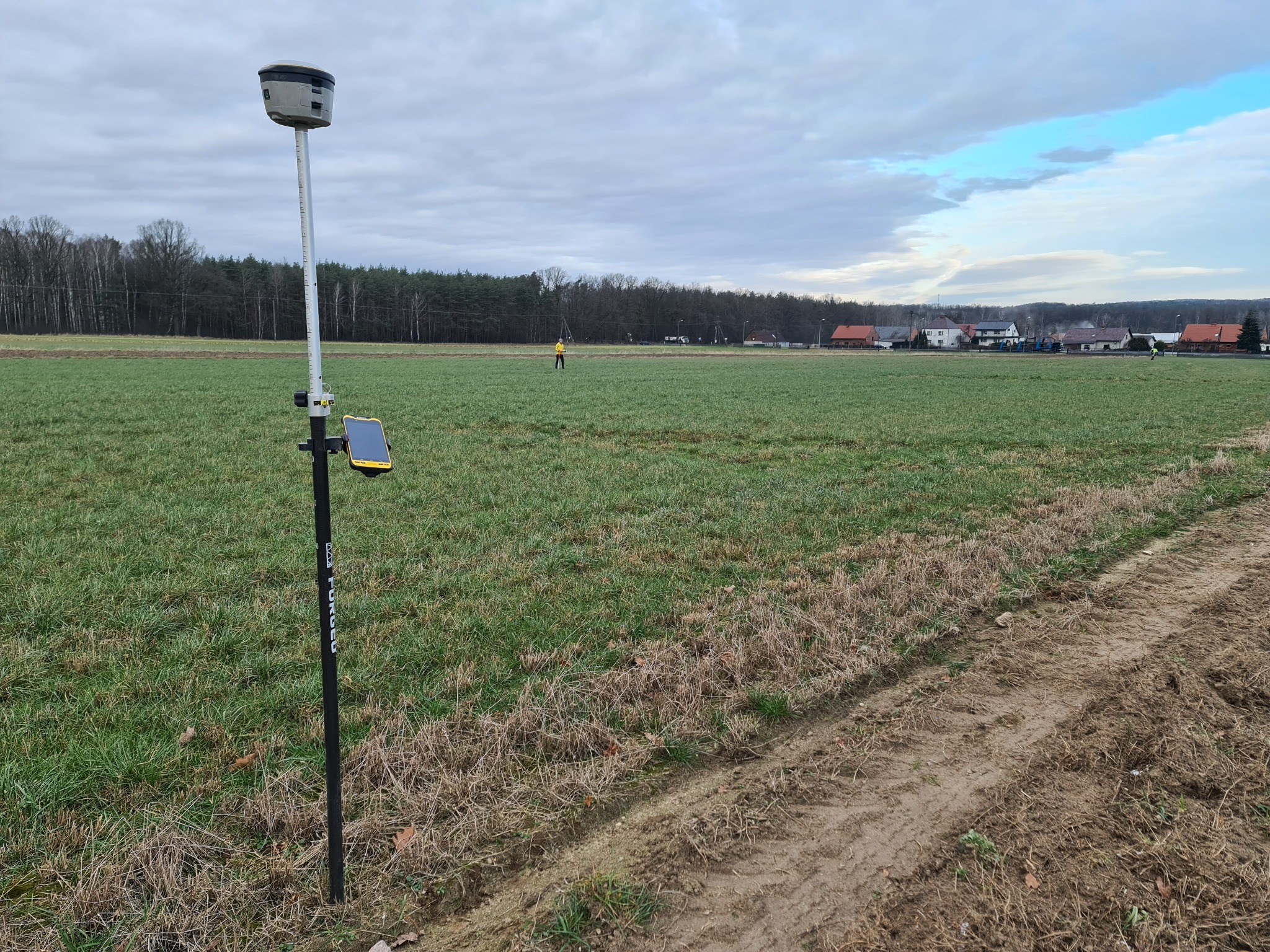 Pomiar geodezyjny na polu: tyczka geodezyjna z odbiornikiem GPS i żółtym smartfonem, w tle pracownik w żółtej kamizelce i zabudowania wiejskie pod pochmurnym niebem.