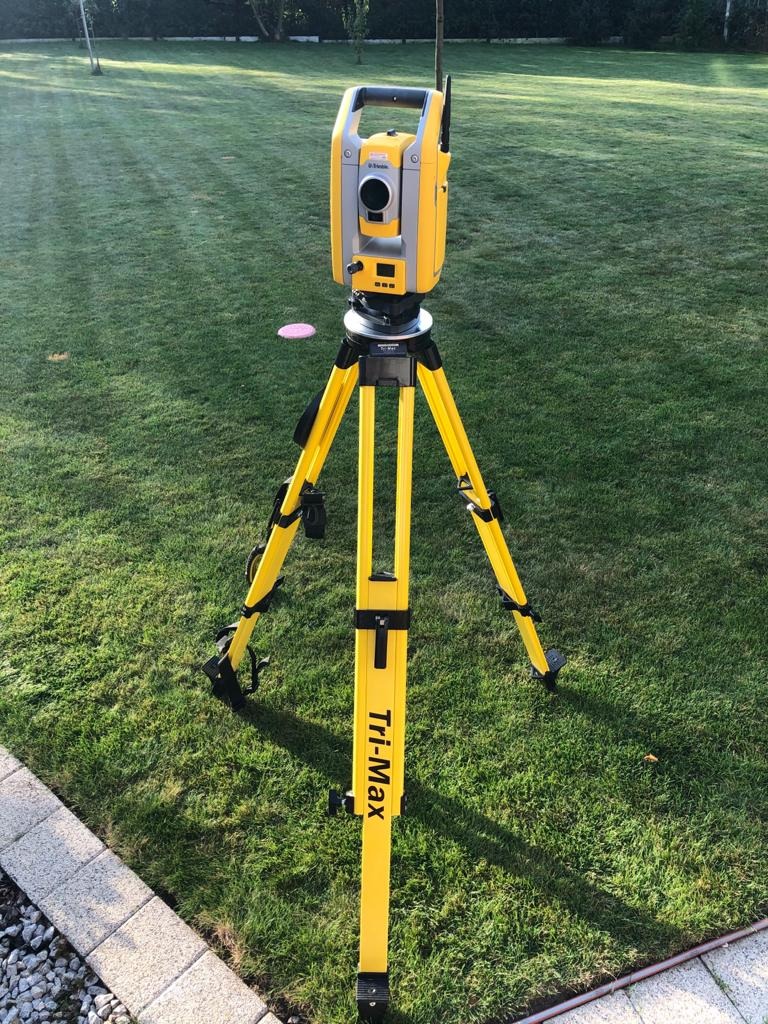 Żółty teodolit Tri-Max ustawiony na trawniku, widok z bliska, w tle drzewa.