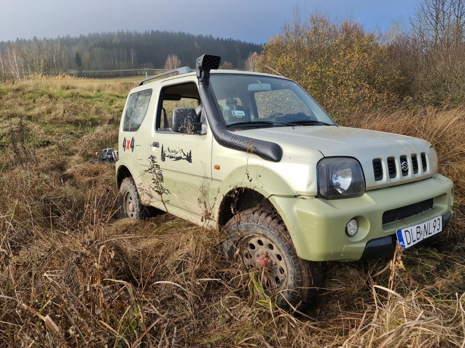 Jasnozielony samochód terenowy Suzuki Jimny z napędem 4x4, z naklejką przedstawiającą góry, brodzi przez błotniste, zarośnięte pole w słoneczny dzień.