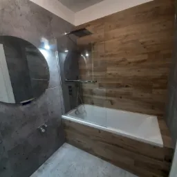 Nowoczesna łazienka z wanną obudowaną panelami imitującymi drewno, szklanym parawanem nawannowym i okrągłym lustrem na szarej, teksturowanej ścianie.