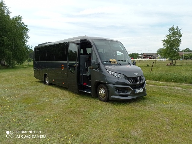 Szary bus marki IVECO Cuby z otwartymi drzwiami, stojący na trawiastym terenie z widocznym budynkiem gospodarczym w tle.