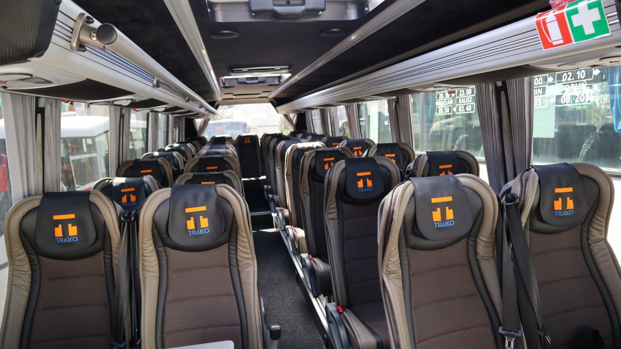 Wnętrze autobusu dalekobieżnego z brązowo-czarnymi fotelami z logo firmy przewozowej TRAKO na zagłówkach, widoczne rozkłady jazdy i oznaczenia bezpieczeństwa.