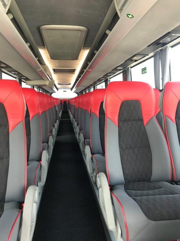 Wnętrze autobusu turystycznego z rzędami szarych foteli z czerwonymi zagłówkami i podłokietnikami, widok z przodu w kierunku tylnej części pojazdu.