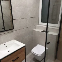 Nowoczesna łazienka z szarymi płytkami, wiszącą toaletą, umywalką z drewnianą szafką i kabiną prysznicową z czarnymi ramami, odbicie prysznica w lustrze.