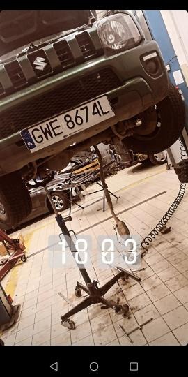 Zielony Suzuki Jimny podniesiony na podnośniku w warsztacie, widoczne podwozie, numer rejestracyjny GWE 8674L, narzędzia i elementy demontowane z pojazdu na podłodze.