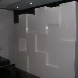 Biała szafa modułowa z błyszczącymi frontami, tworząca geometryczny wzór na ścianie, widoczny fragment czarnego blatu kuchennego i okno z żaluzjami.