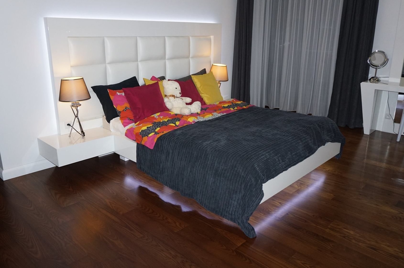 Nowoczesna sypialnia z białym łóżkiem z podświetlanym zagłówkiem i szafkami nocnymi, drewniana podłoga, ciemne zasłony, pluszowy miś na łóżku.