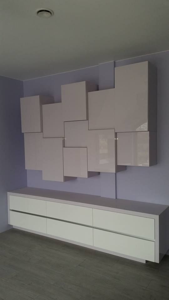 Biała szafka RTV z sześcioma szufladami i asymetrycznie rozmieszczone, lakierowane szafki wiszące na fioletowej ścianie, szare panele podłogowe.