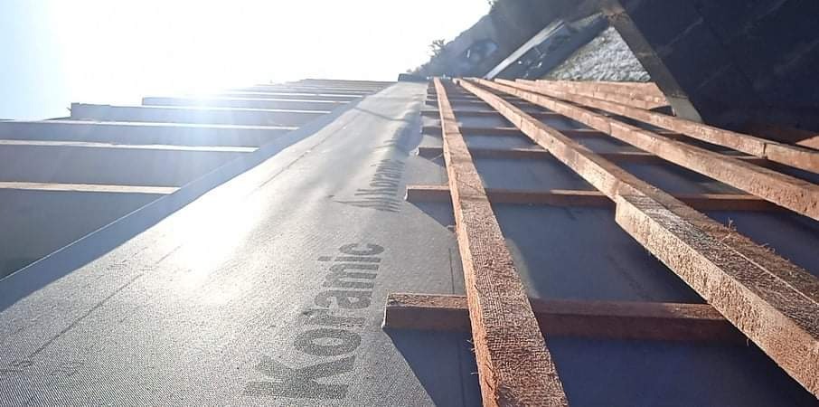 Widok z dołu na konstrukcję dachu w trakcie budowy, z widoczną membraną dachową Koramic i drewnianymi łatami, oświetlone jasnym słońcem.