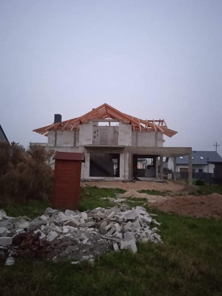 Surowy stan budowy domu jednorodzinnego z widoczną konstrukcją dachu i stertą gruzu na pierwszym planie, pochmurne niebo w tle.