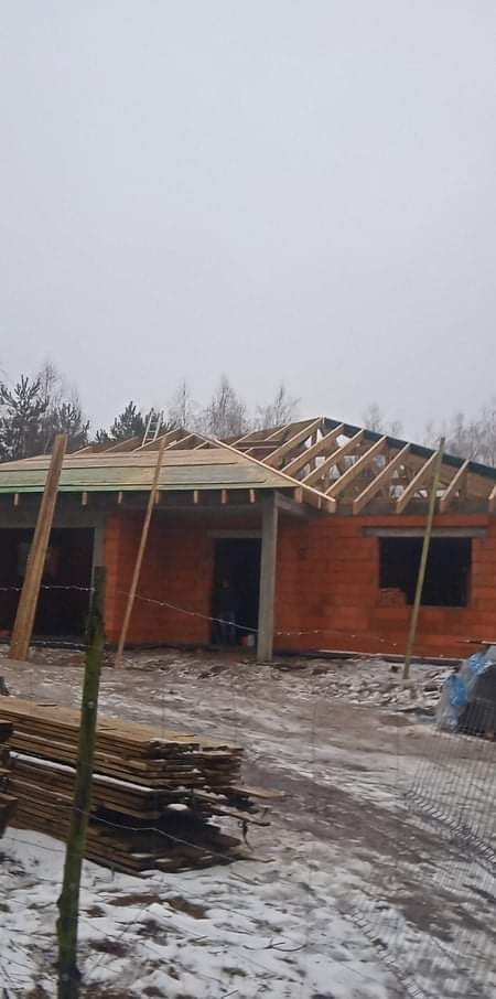 Budowa domu jednorodzinnego w trakcie montażu więźby dachowej, widoczne drewniane elementy konstrukcyjne i fragment muru z czerwonej cegły, w tle las i zimowy krajobraz.