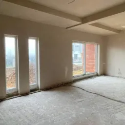 Wnętrze pokoju po nałożeniu tynków, z widocznymi oknami i surowym wykończeniem podłogi. Widoczne elementy instalacji elektrycznej.