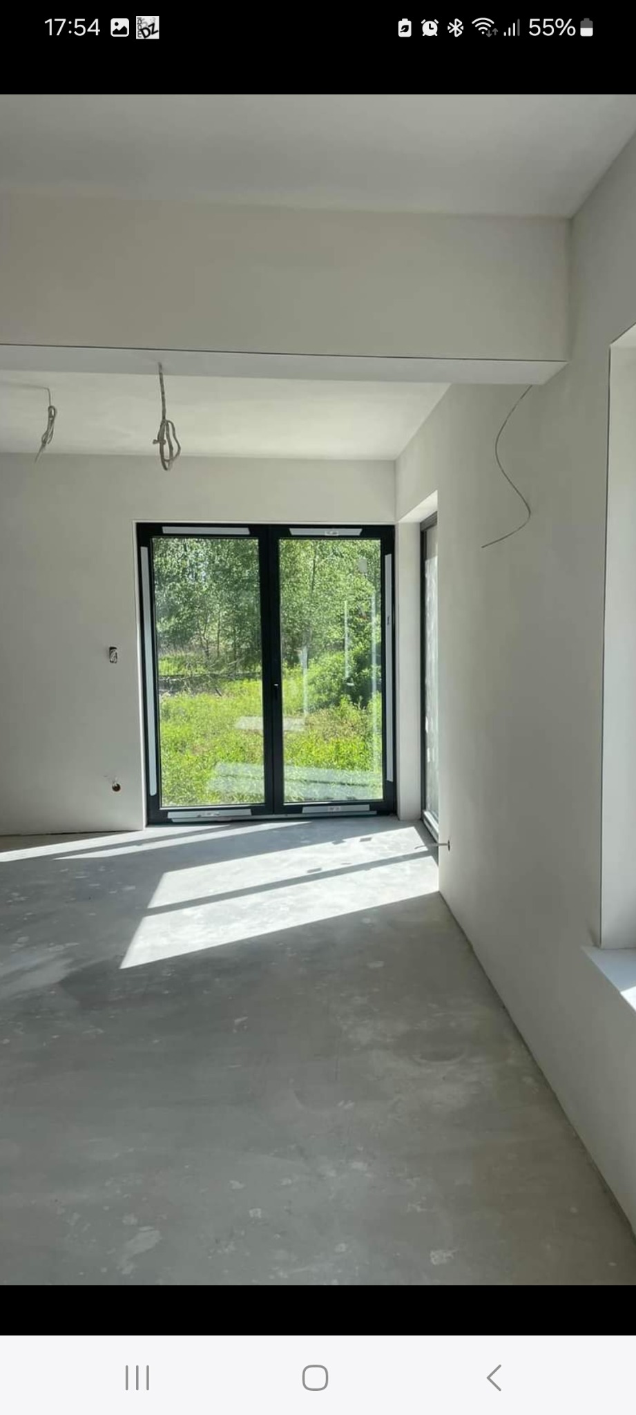 Surowe wnętrze po tynkowaniu maszynowym, widoczne świeżo otynkowane ściany, betonowa posadzka i czarne ramy okienne z widokiem na zieleń na zewnątrz, z wiszącymi kablami elektrycznymi