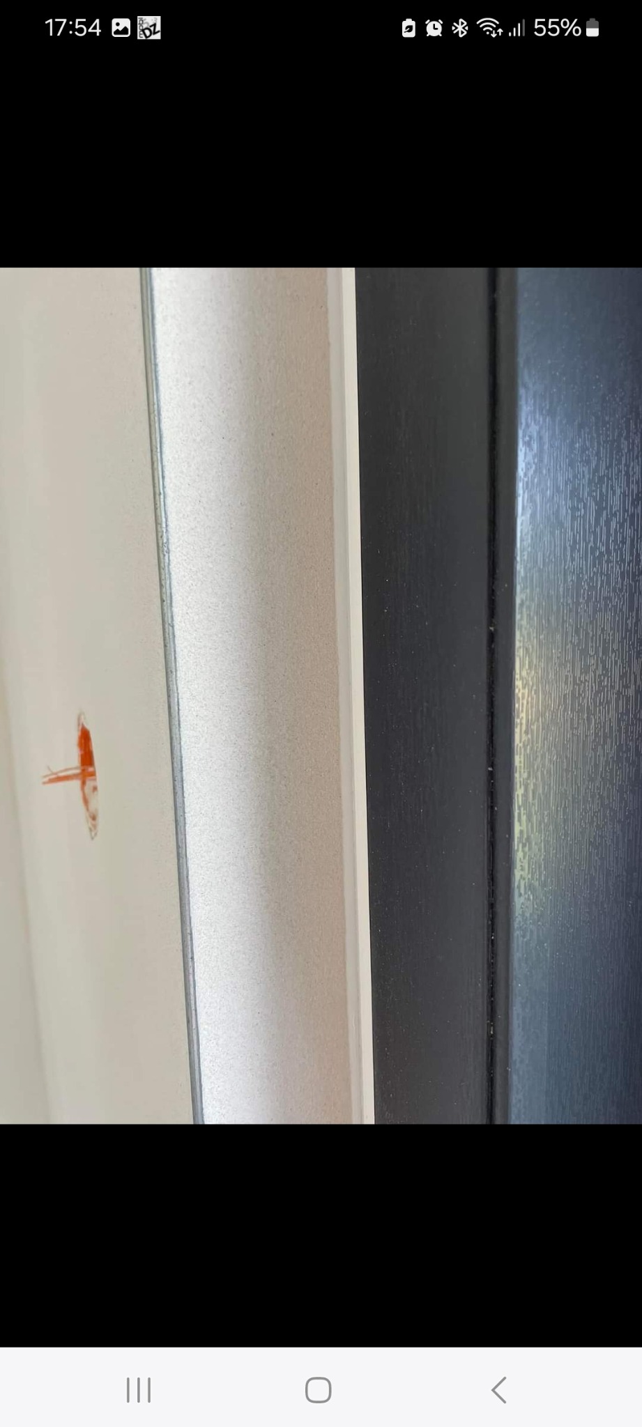 Fragment ściany z niedoskonałością tynku (niewielki pomarańczowy ślad) sąsiadującej z ciemną framugą drzwi.