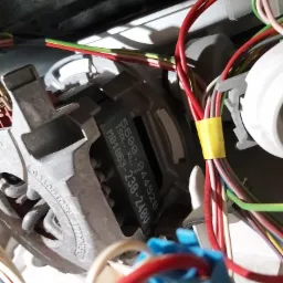 Silnik elektryczny z widocznymi uzwojeniami i tabliczką znamionową, otoczony wiązką kolorowych przewodów z żółtą taśmą izolacyjną, prawdopodobnie element pralki.