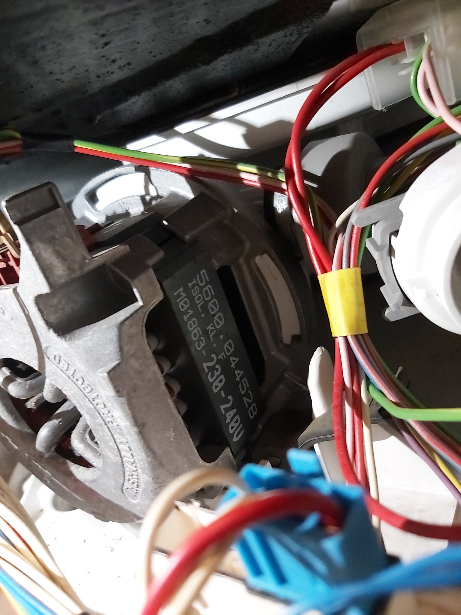 Silnik elektryczny z widocznymi uzwojeniami i tabliczką znamionową, otoczony wiązką kolorowych przewodów z żółtą taśmą izolacyjną, prawdopodobnie element pralki.