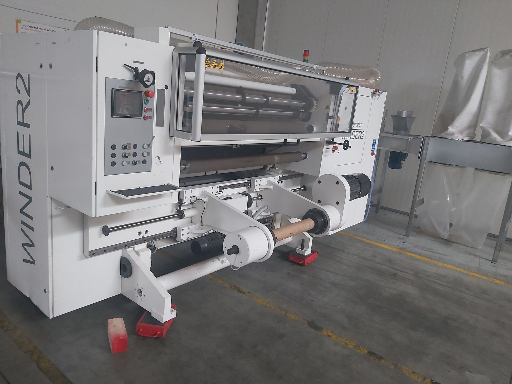Biała maszyna przemysłowa WINDEER2 z widocznymi panelami sterowania, osłonami bezpieczeństwa i rolką materiału w uchwytach, na tle szarej posadzki z czerwonymi podkładkami pod nogami.