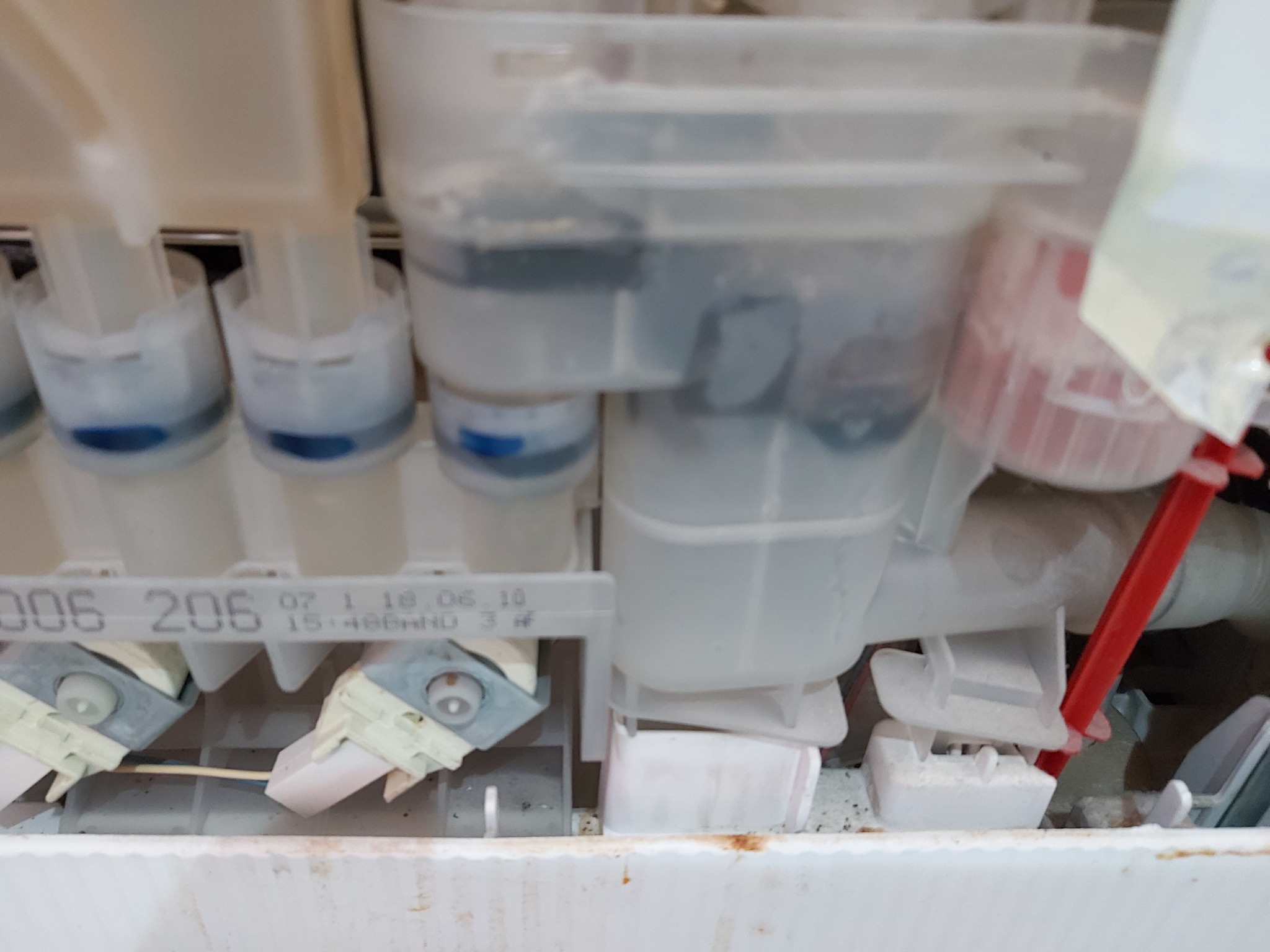 Wnętrze zmywarki w trakcie naprawy w Krakowie, widoczne elementy układu doprowadzania wody i dozowania detergentu, z numerami seryjnymi i datami produkcji na plastikowych komponentach.