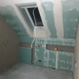 Poddasze w trakcie wykańczania: zamontowane płyty kartonowo-gipsowe na ścianach i suficie, widoczne szpachlowane łączenia, okno dachowe, wystające przewody elektryczne.