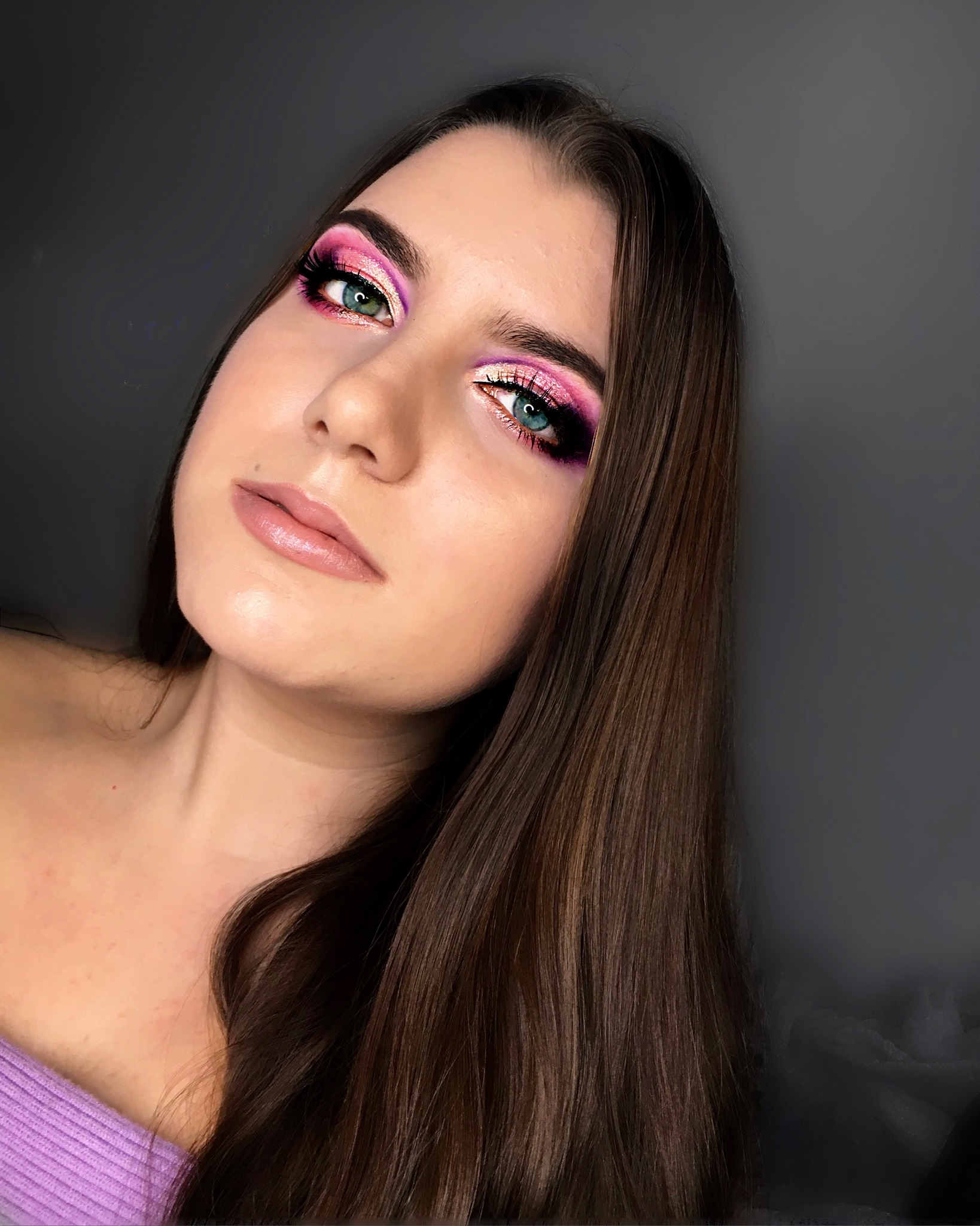 Portret kobiety z intensywnym, kolorowym makijażem oczu w odcieniach różu, fioletu i złota, z błyszczącymi ustami i długimi, brązowymi włosami na tle ciemnoszarej ściany.