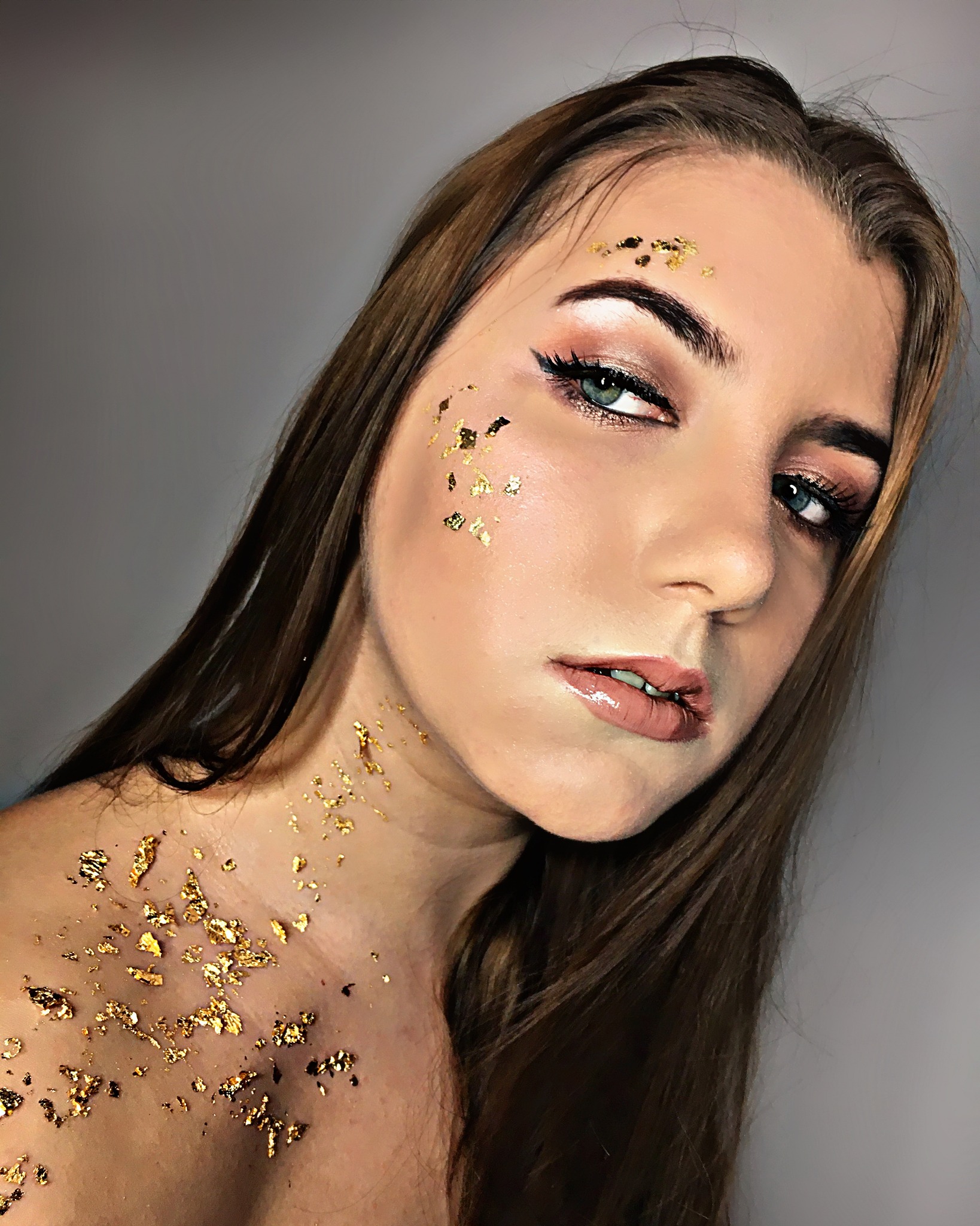 Portret kobiety z wyrazistym makijażem oczu i złotymi płatkami na twarzy oraz dekolcie, podkreślającymi artystyczny charakter stylizacji.