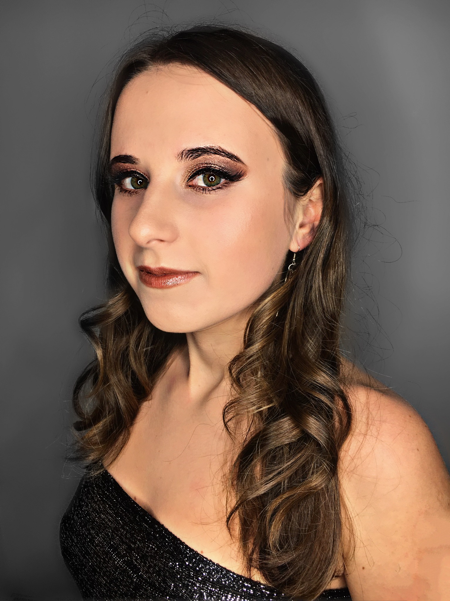 Portret kobiety z profesjonalnym makijażem oczu w odcieniach brązu i błyszczącą pomadką, z delikatnymi kręconymi włosami i subtelną biżuterią na uszach, na jednolitym szarym tle.