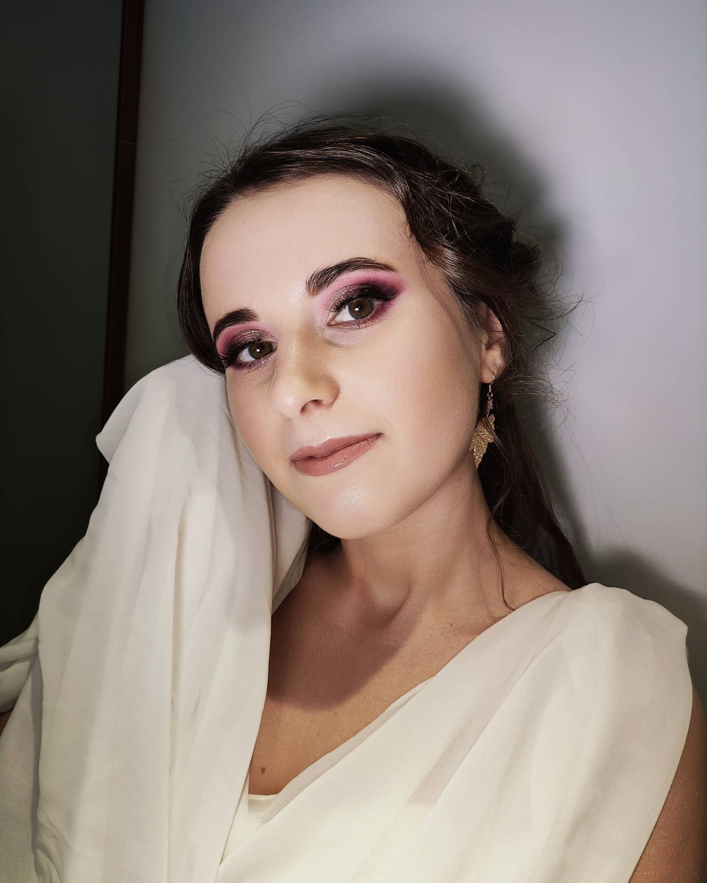 Portret kobiety z wyrazistym, różowo-fioletowym cieniowaniem powiek, podkreślonymi brwiami i błyszczącymi ustami w odcieniu nude, pozującej z kremową tkaniną drapowaną na ramieniu oraz złotymi...