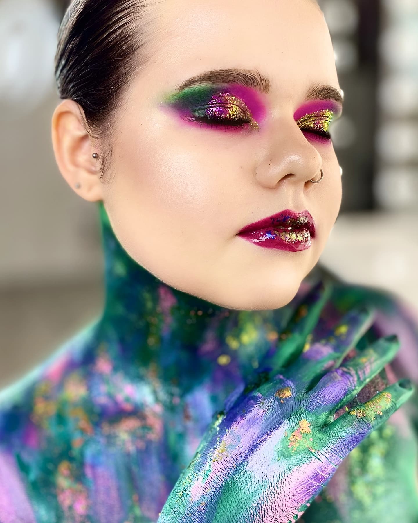 Portret kobiety z artystycznym makijażem oczu w odcieniach fioletu, zieleni i złota oraz pomalowanym ciałem w abstrakcyjne wzory, pierścień w nosie i kolczyk w uchu.