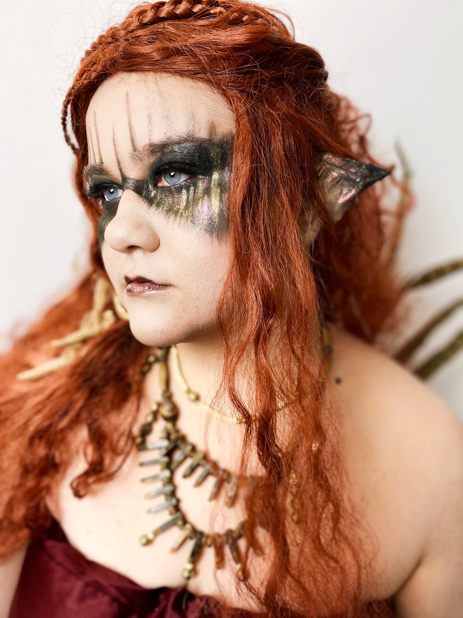 Portret kobiety z rudymi włosami i fantazyjnym makijażem: ciemne cienie z brokatem, pionowe paski na czole, szpiczaste uszy i ozdobny naszyjnik, stylizacja fantasy.
