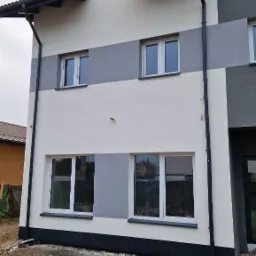 Dwukondygnacyjny dom z ocieploną elewacją w odcieniach bieli i szarości, z ciemnymi rynnami i widocznym fragmentem dachu sąsiedniego budynku, teren wokół domu jeszcze niezagospodarowany.