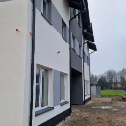 Nowoczesny budynek wielorodzinny z elewacją w odcieniach szarości i bieli, widoczne orynnowanie i fragment terenu przed budynkiem po pracach ziemnych.