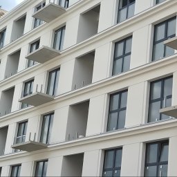 Przembud Przemysław Tokarzewski - Nowoczesny budynek z jasną elewacją i ciemnymi oknami, z wystającymi betonowymi płytami balkonowymi. Widoczne rusztowanie po prawej stronie, sugerujące trwające prace wykończeniowe.