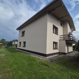 Przembud Przemysław Tokarzewski - Dom jednorodzinny z nową, jasnobeżową elewacją i ciemnobrązowym cokołem, brązowe okna i balustrada balkonu, na którym stoi osoba. W tle częściowo zachmurzone niebo i zielony trawnik.