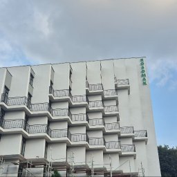 Przembud Przemysław Tokarzewski - Nowoczesny budynek z białą elewacją i kaskadowo ułożonymi balkonami z czarnymi balustradami. Na prawej fasadzie zielony napis 'ROSOMAK'. W dolnej części widoczne rusztowania i pracownik na nich.