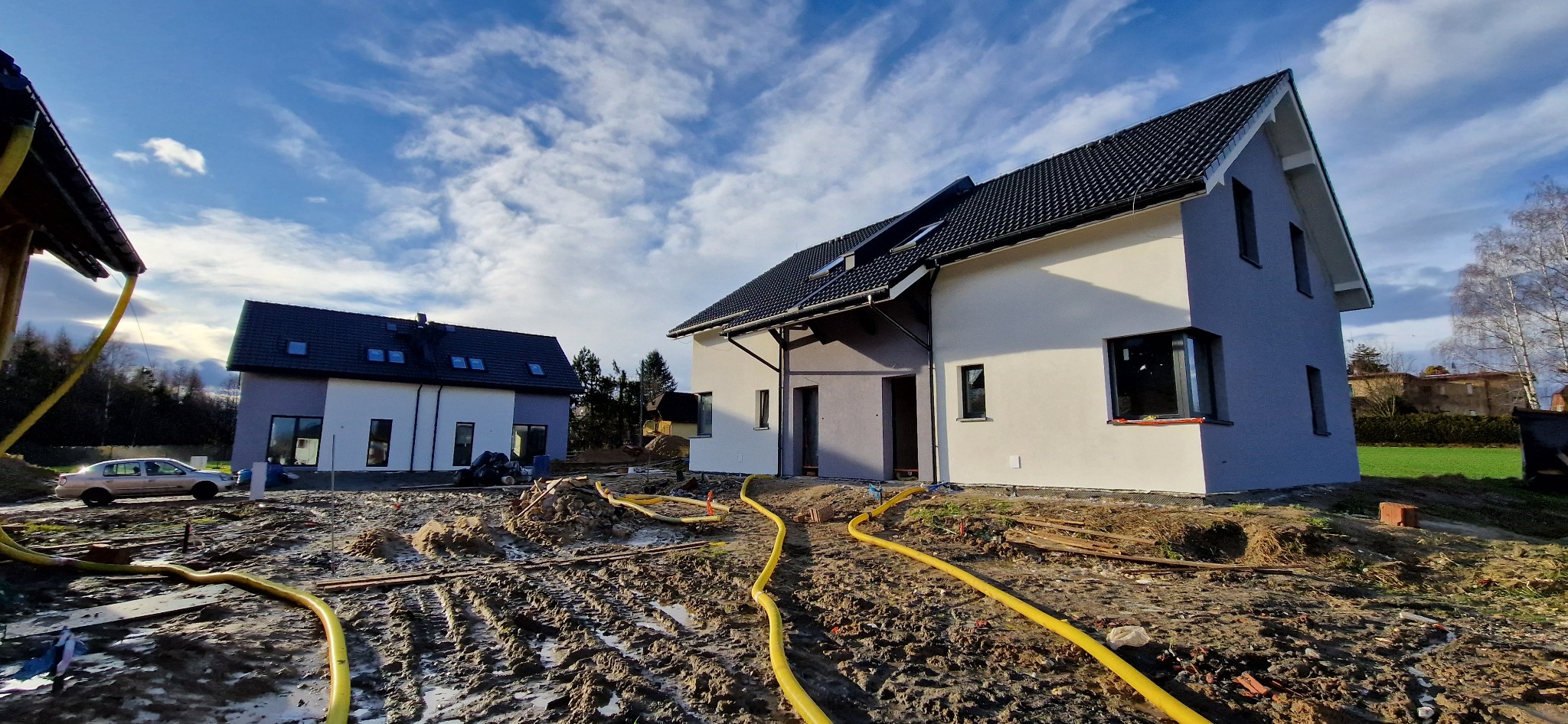 Dwa domy jednorodzinne w trakcie budowy, elewacja częściowo pomalowana na biało i szaro, teren wokół rozkopany, widoczne żółte węże, błękitne niebo z chmurami w tle.