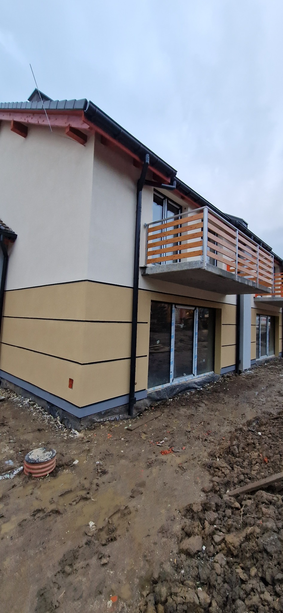 Dwukondygnacyjny dom z wykończoną elewacją w dwóch odcieniach beżu, z balkonami z drewnianą balustradą, widok na fragment budynku z ziemistym terenem wokół.