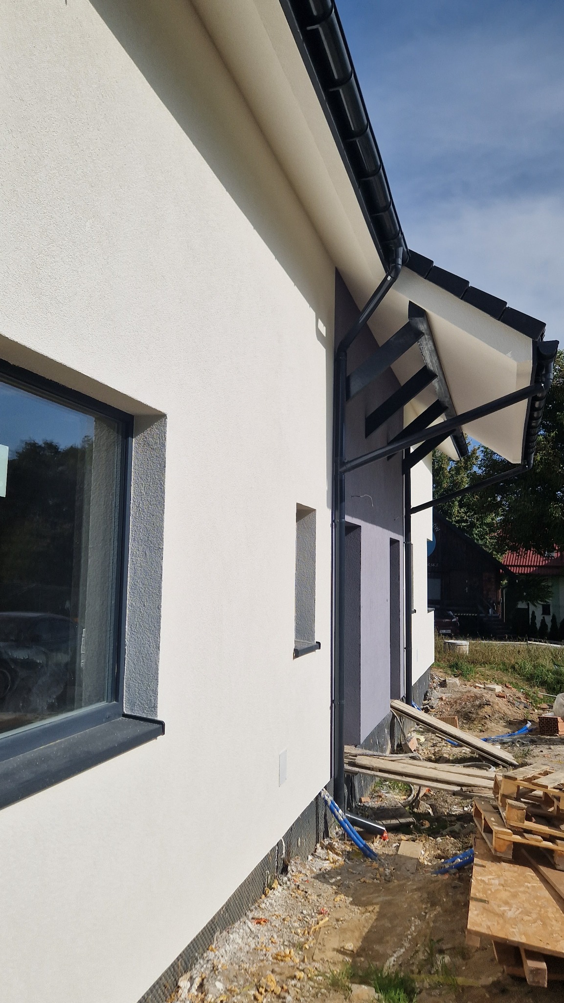 Fragment elewacji domu jednorodzinnego z widocznym oknem, rynną i zadaszeniem nad wejściem, z wykończonym tynkiem strukturalnym w jasnym odcieniu i częściowo pomalowaną ścianą w kolorze szarym...