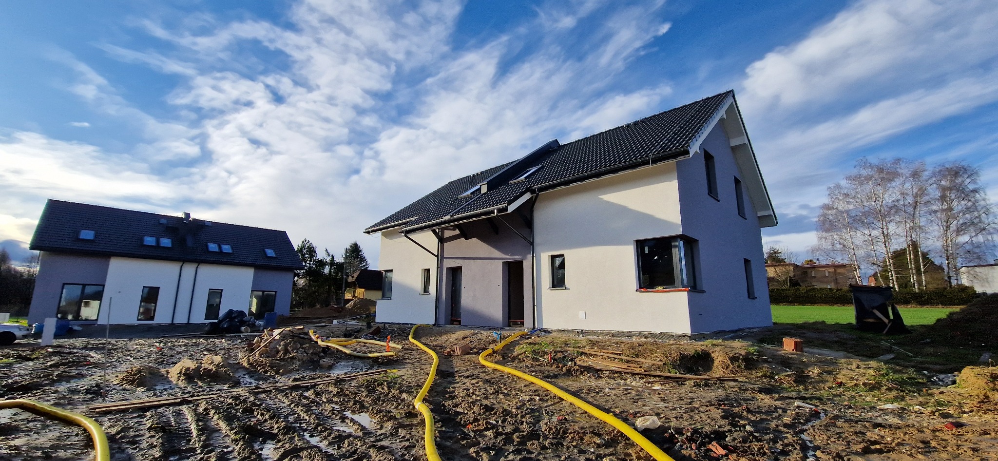 Dwukondygnacyjny dom jednorodzinny z szarą i białą elewacją, czarnym dachem z oknami dachowymi i widocznymi żółtymi wężami na błotnistym terenie wokół budynku.