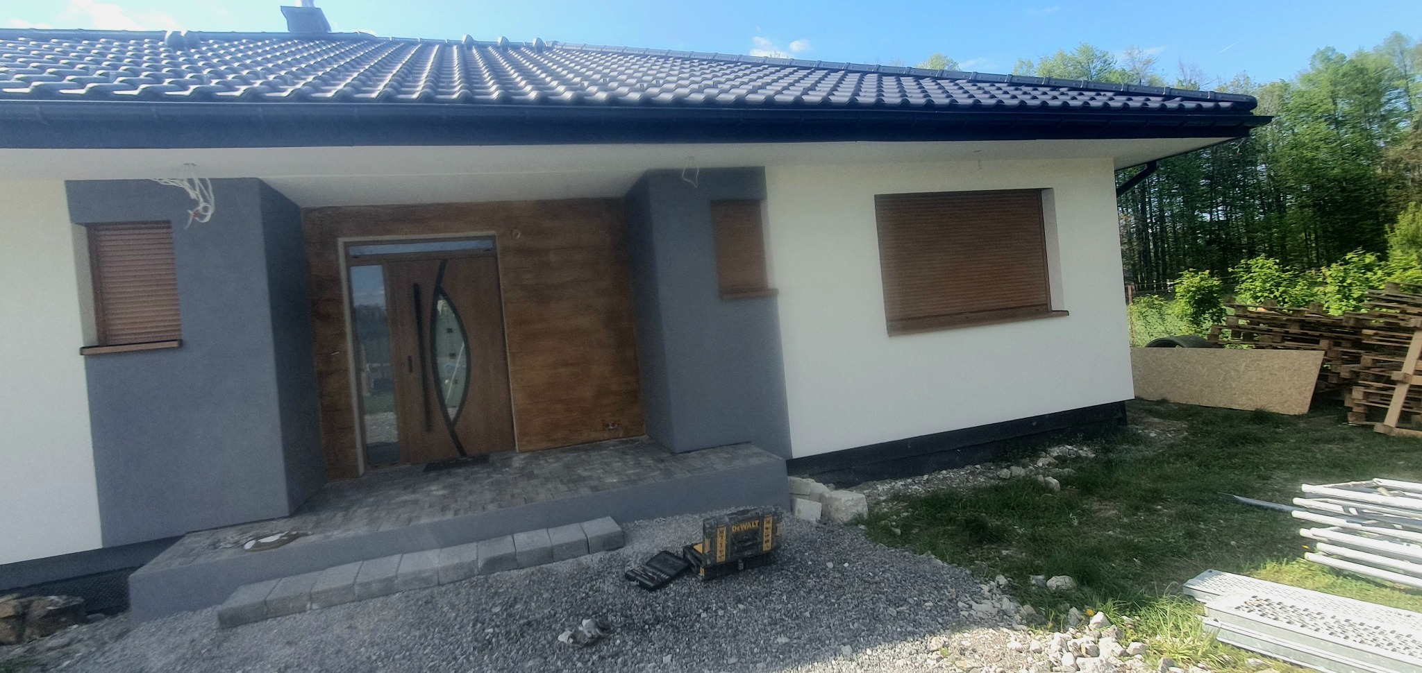 Nowoczesny dom jednorodzinny z szarą elewacją i drewnopodobnym wykończeniem przy wejściu, widoczne rolety zewnętrzne, teren wokół domu w trakcie zagospodarowania, na pierwszym planie skrzynka...