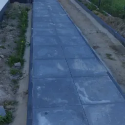 Długa, prosta ścieżka z ciemnoszarych, kwadratowych płyt brukowych prowadząca wzdłuż białej ściany budynku i ogrodzenia z siatki, z widocznym klimatyzatorem na ścianie i fragmentem trawnika.