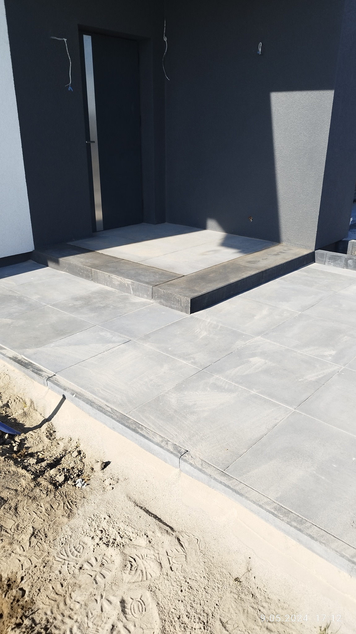 Wykończone wejście do domu z szarej kostki brukowej, widoczne ślady prac ziemnych wzdłuż krawędzi, szara elewacja budynku w tle.