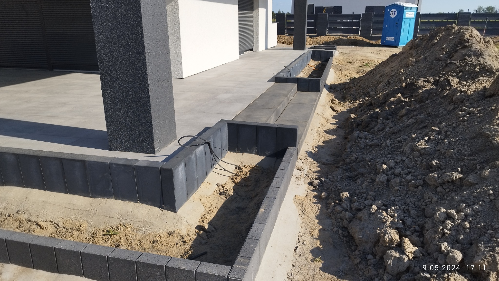 Przygotowanie podłoża pod obrzeża z kostki brukowej, widoczne kable i wykopana ziemia, w tle dom z szarą elewacją i przenośna toaleta.