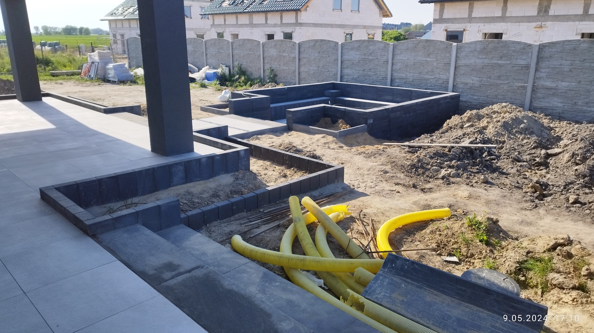 Budowa tarasu z ciemnej kostki brukowej, widoczne żółte rury instalacyjne i sterta piasku, dom w tle.
