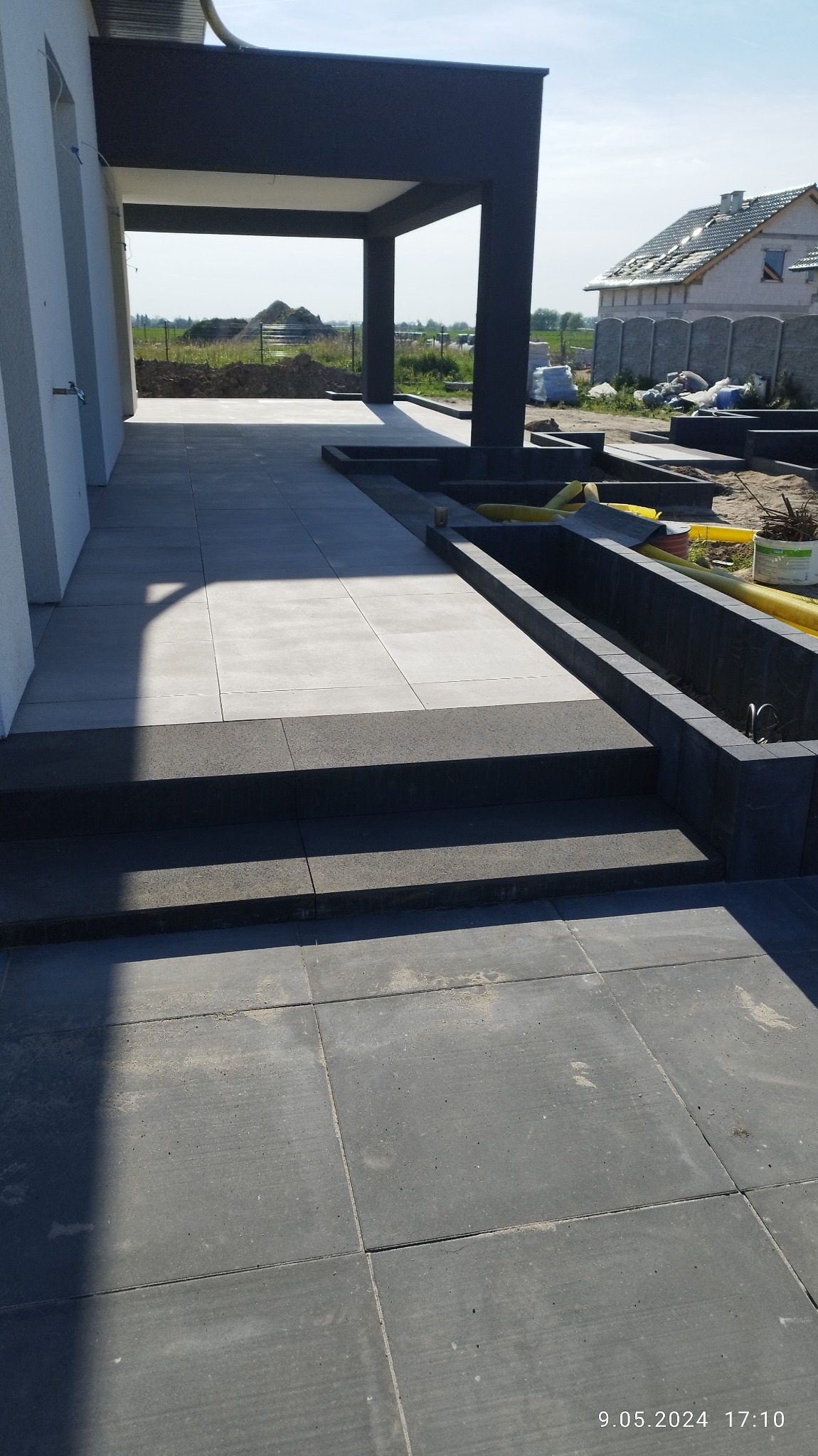 Nowoczesny taras z szarych płyt betonowych, schody z ciemniejszej kostki, widoczne elementy instalacji nawadniającej w przygotowanych rabatach, w tle budowa domu.